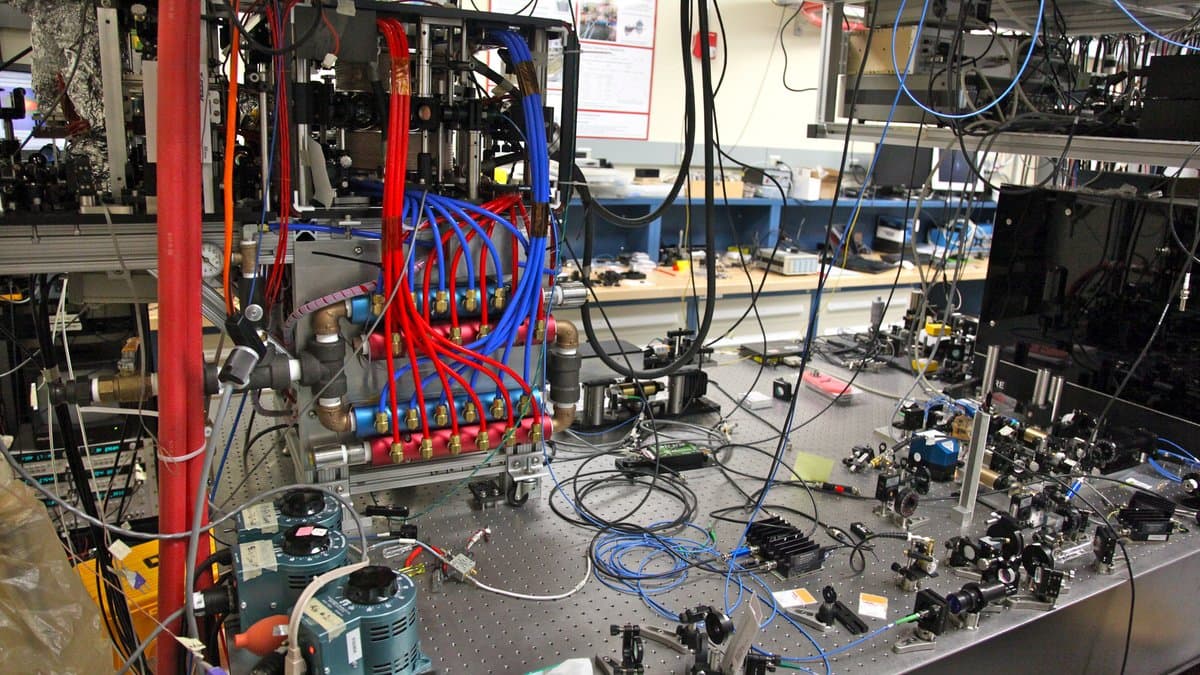 An optical table in a physics laboratory, crowded with mirrors, lenses and cabling around a vacuum chamber used to trap laser-cooled atoms.