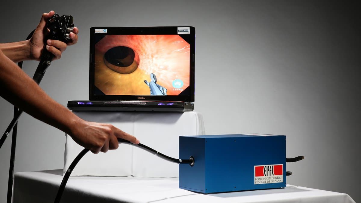 Two hands steer a flexible endoscope on a benchtop colonoscopy training simulator, with a laptop screen showing the simulated view from the scope tip.