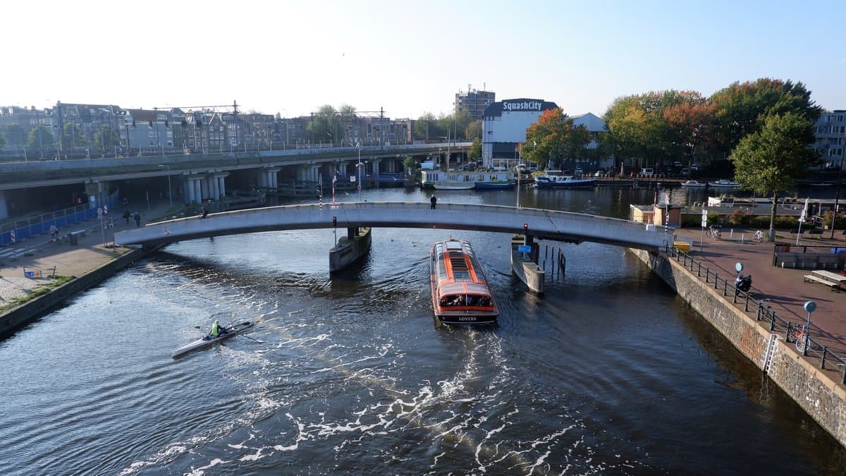 An Amsterdam canal seen from above, with a line of air bubbles and foam crossing the water beside a bridge and a passing tour boat.
