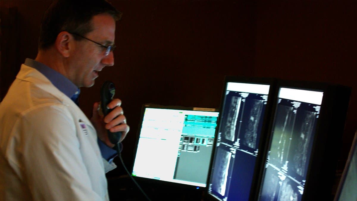 A radiologist in a white coat dictates a report while reading cross-sectional scans on two display monitors in a darkened reading room.