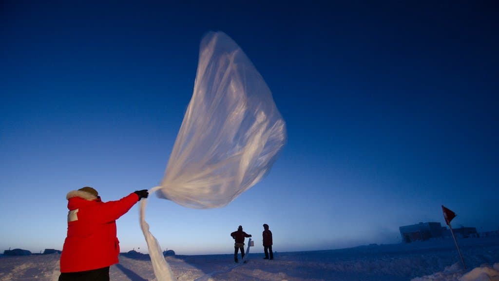 Two researchers on a snowfield in Antarctica release a large translucent balloon into a deep blue twilight sky, with station buildings on the horizon.