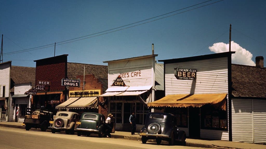 A small-town main street in Cascade, Idaho, in 1941, with storefront awnings pulled out over the sidewalk, parked cars of the period and a man standing in the shade under a clear sky.