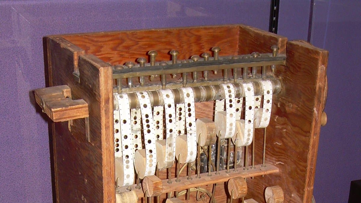 A wooden cabinet holding rows of punched steel chains and brass gears, an early machine for number theory calculations, inside a museum display case.