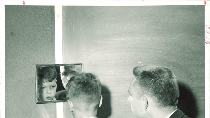 Black and white photograph of a boy facing a small wall mirror beside a clinician during a speech therapy session in a 1950s school room, the boy's face reflected in the mirror.