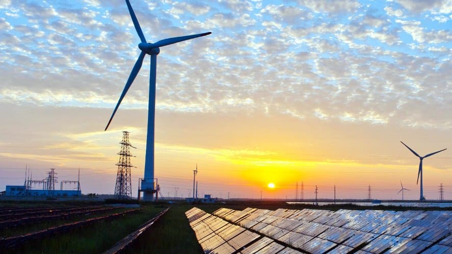 A large wind turbine stands beside rows of solar panels, with electricity pylons and a substation behind them under a cloud-streaked sunset sky.