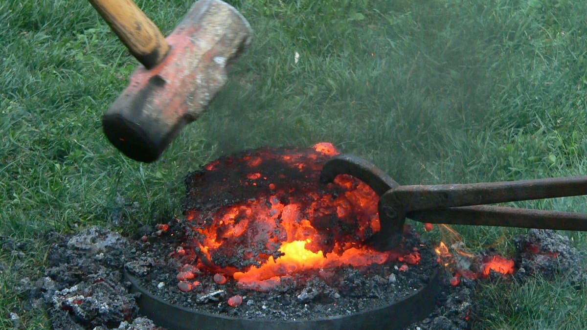 A glowing orange mass of freshly smelted iron on the ground, struck with a sledgehammer and gripped with long tongs