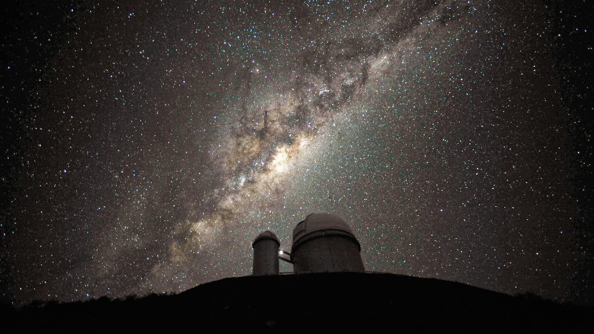 Two telescope domes on a dark ridge at La Silla Observatory, silhouetted against the bright band and central bulge of the Milky Way.