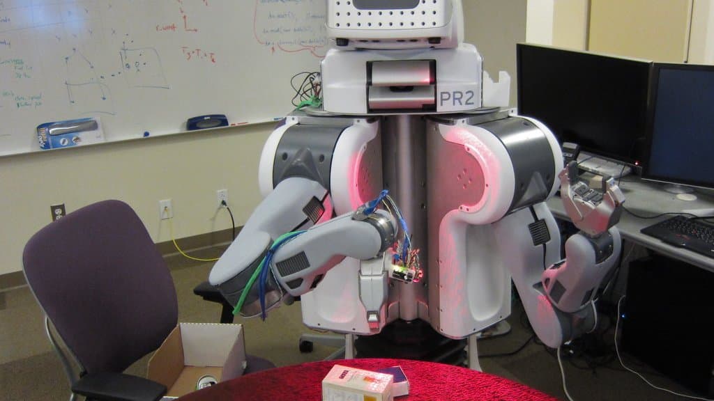 A two-armed white research robot stands at a laboratory table, its grippers extended over boxes and cans, with a whiteboard and computer monitors behind it.