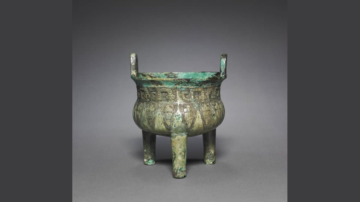 A green-patinated bronze tripod cauldron with two upright handles and a band of cast animal-mask ornament, photographed against a plain gray background.