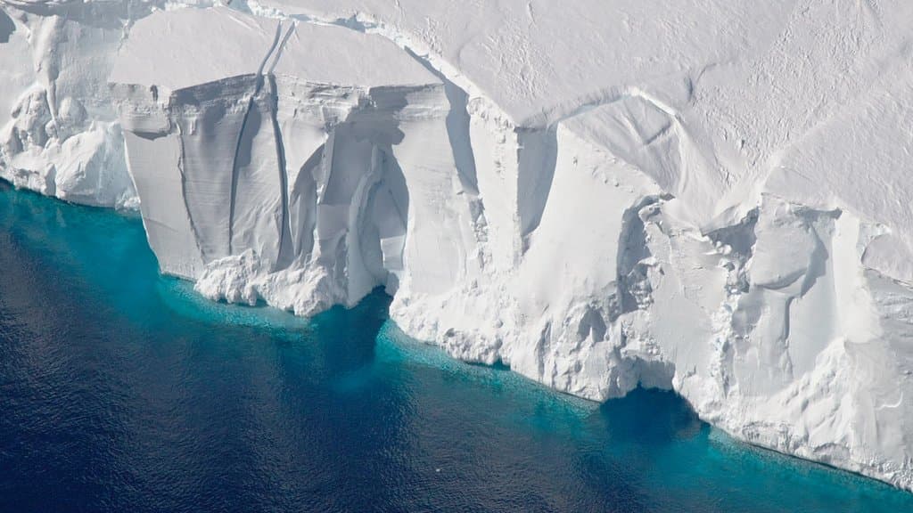 Aerial view of a tall white ice cliff at the front of the Getz Ice Shelf meeting turquoise ocean water.