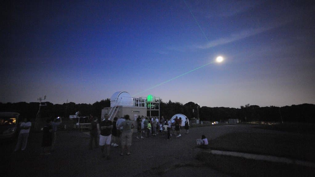 A thin green laser beam rises from a small domed observatory at night and passes close to a bright Moon while a group of visitors watches from the parking area.