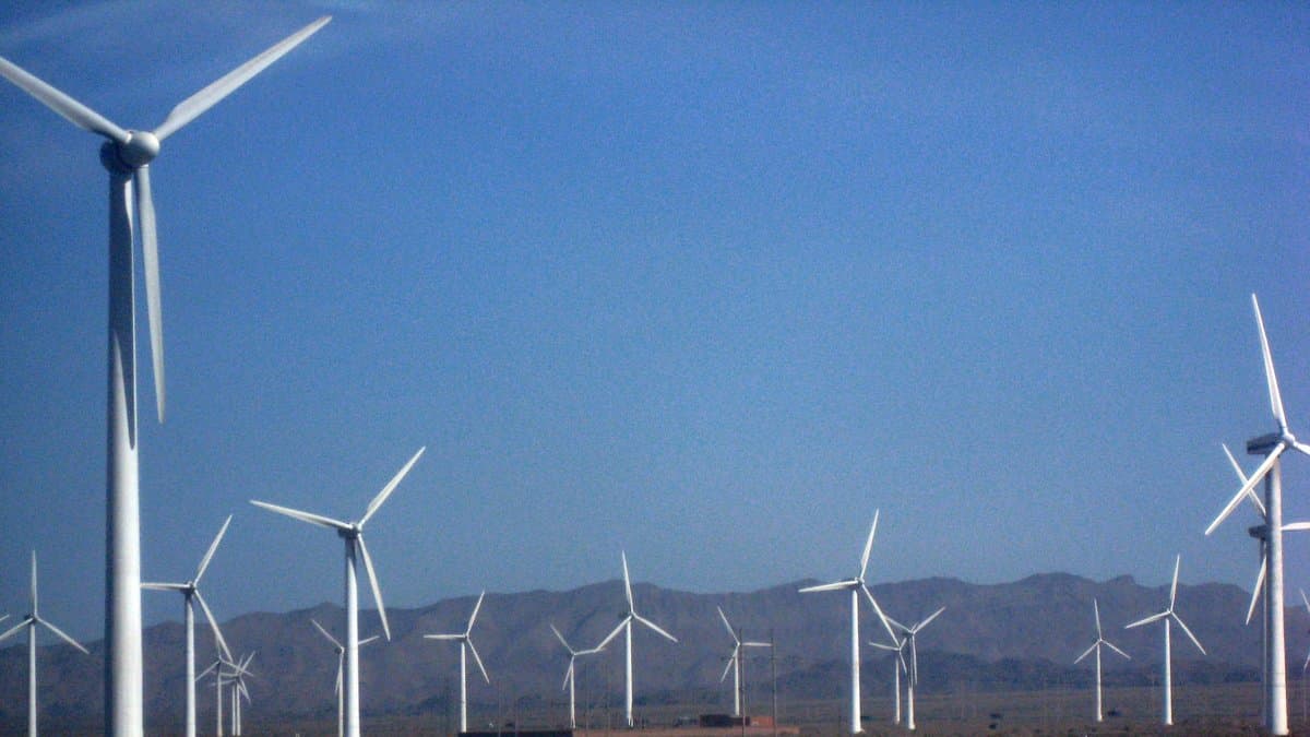 Rows of wind turbines on a gravel desert plain in Xinjiang, China, with a long mountain ridge behind them.