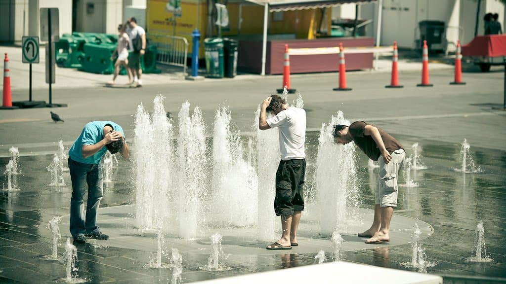 Three people bend under the jets of a public plaza fountain to cool their heads during hot summer weather in Montreal.