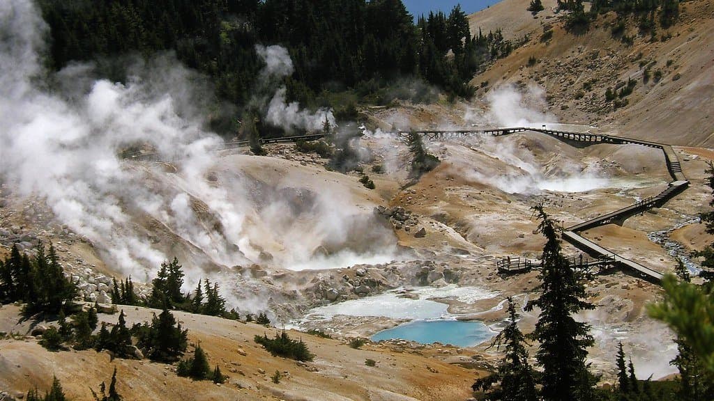 Steam rising from fumaroles across a pale, sulfur-stained basin with a turquoise hot pool and a wooden boardwalk, ringed by conifers and distant ridges