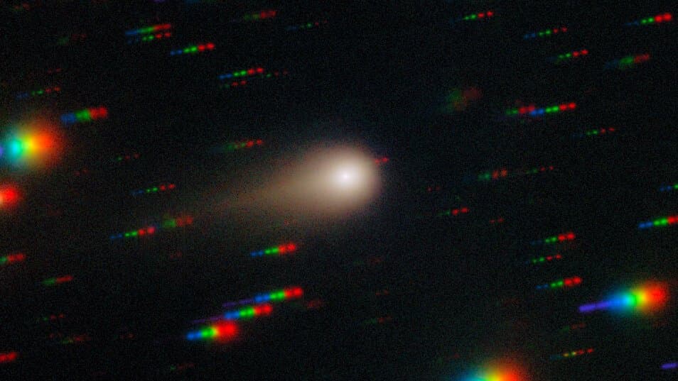 Interstellar comet 3I/ATLAS as a bright fuzzy nucleus with a short tail, held steady while background stars streak into short red, green and blue dashes.