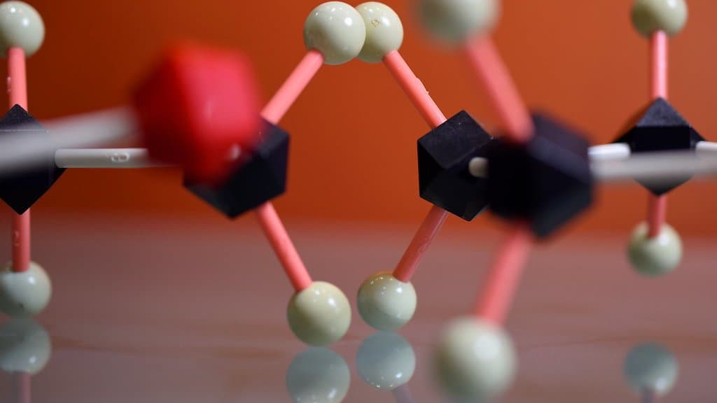 Close-up photograph of a plastic ball-and-stick molecular model: cream and red spheres joined by pink rods to black connectors, against an orange background.