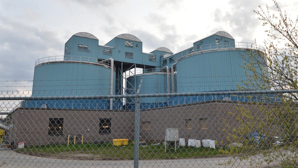Blue cylindrical digester tanks and a brick service building at a municipal wastewater treatment plant, seen through a chain-link fence.