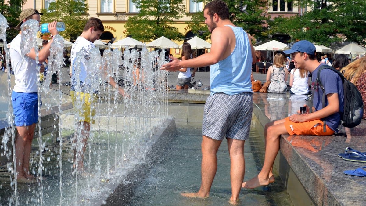 People stand and sit in the jets of a public fountain on a city square in Krakow, Poland, with tenement houses and market umbrellas behind them.