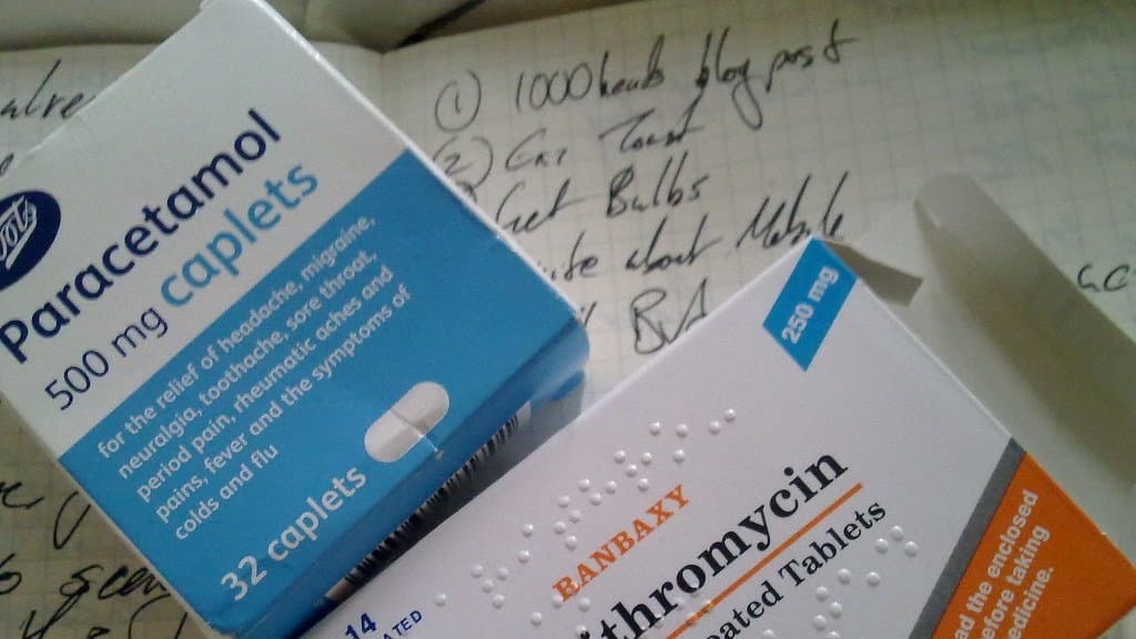 A blue and white carton of paracetamol 500 mg caplets lying on a notebook beside a carton of clarithromycin tablets.