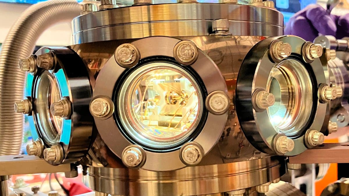 A stainless steel ultra-high-vacuum chamber with bolted circular viewports, the central window glowing, housing a linear Paul ion trap.