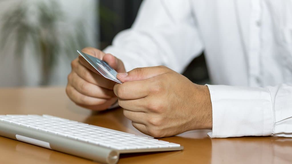 A person in a white shirt sits at a desk holding a payment card in both hands, with a computer keyboard in the foreground.