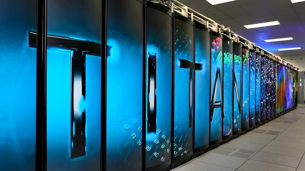 A long row of black supercomputer cabinets in a data center aisle, lit blue and printed with the word TITAN and scientific imagery, receding toward the back of the room.