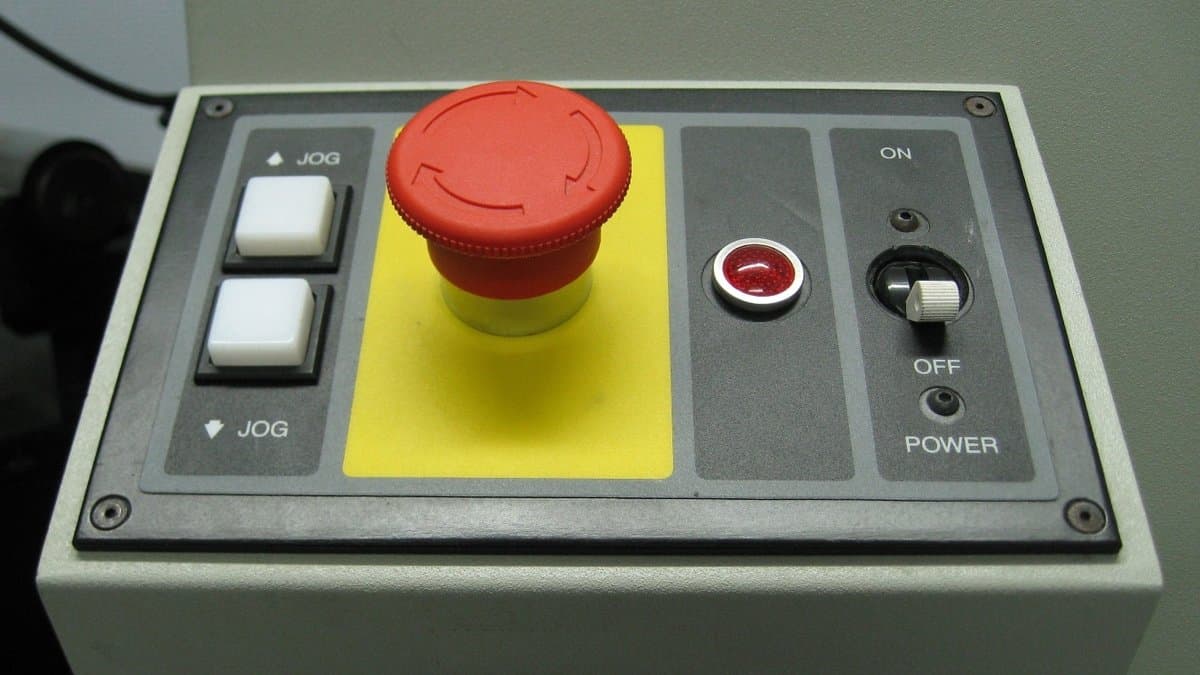A red mushroom-shaped emergency stop button on a gray machine control panel, beside a toggle switch labeled ON and OFF.