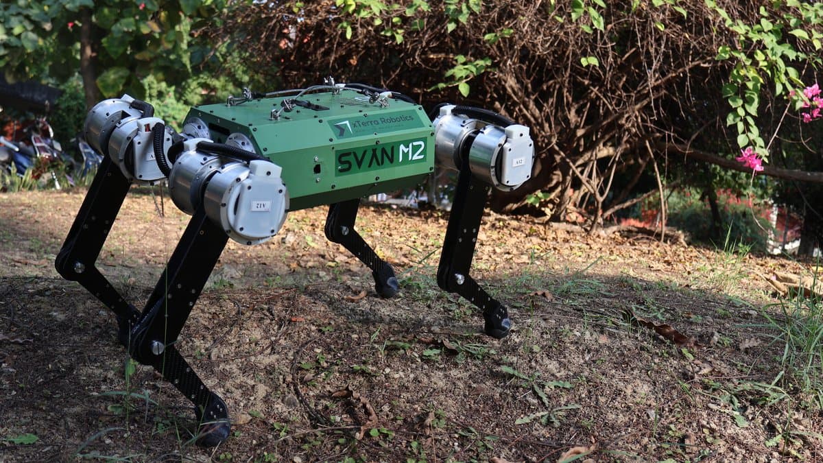 A green and black four-legged robot labeled SVAN M2 standing on rough ground in front of trees.