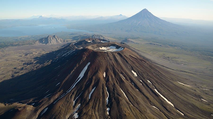 Aerial view of Krasheninnikov volcano in Kamchatka, a broad dark cone with a snow-streaked summit crater, with a lake and a tall pointed volcanic peak in the distance.