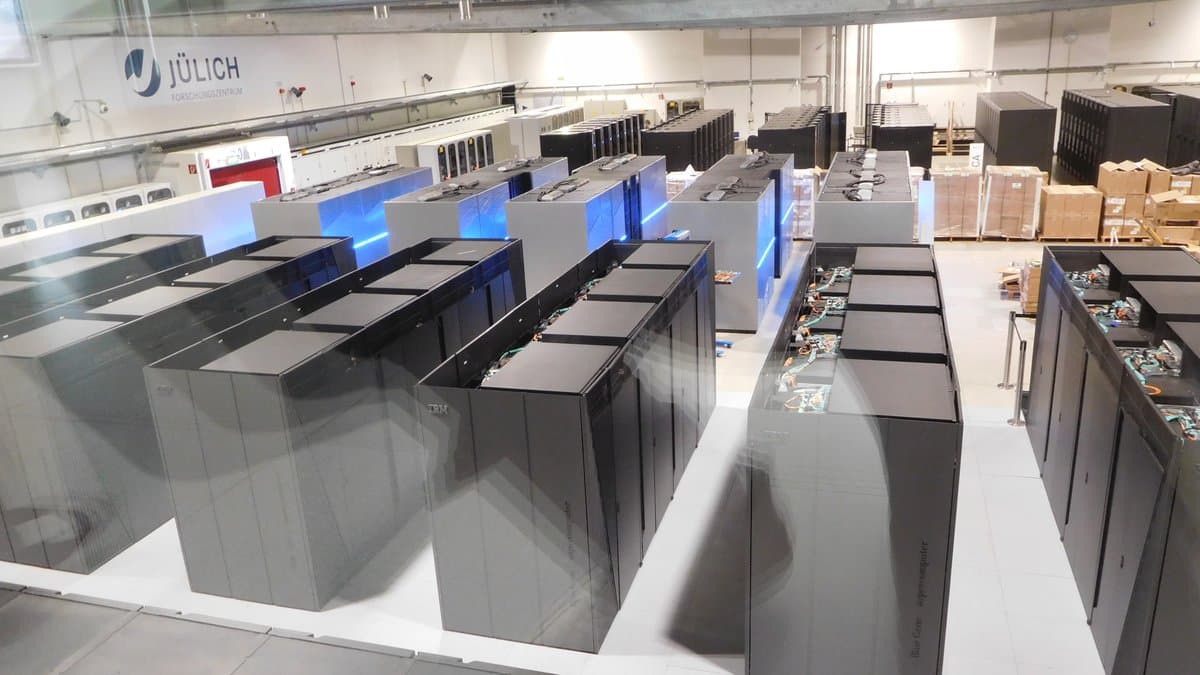 Rows of tall black supercomputer cabinets in a machine hall, with the Forschungszentrum Jülich logo on the wall behind them.