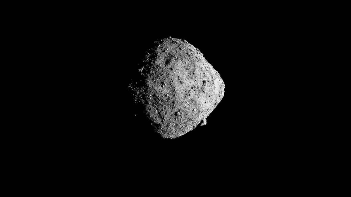 The asteroid Bennu, a dark diamond-shaped rubble pile covered in boulders, lit from one side against black space.