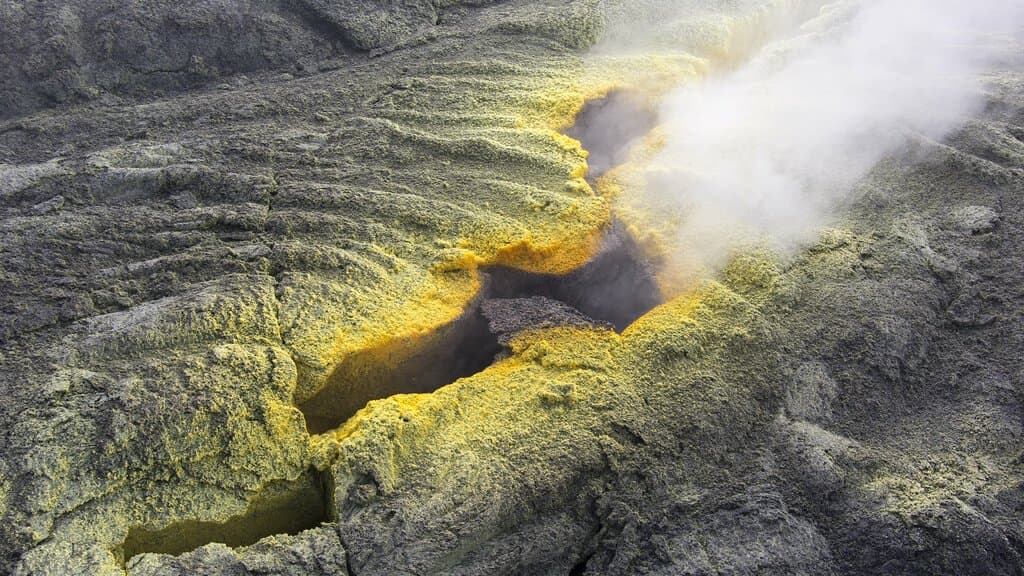 Bright yellow elemental sulfur encrusting the rim of a steaming fumarole vent in dark volcanic rock.