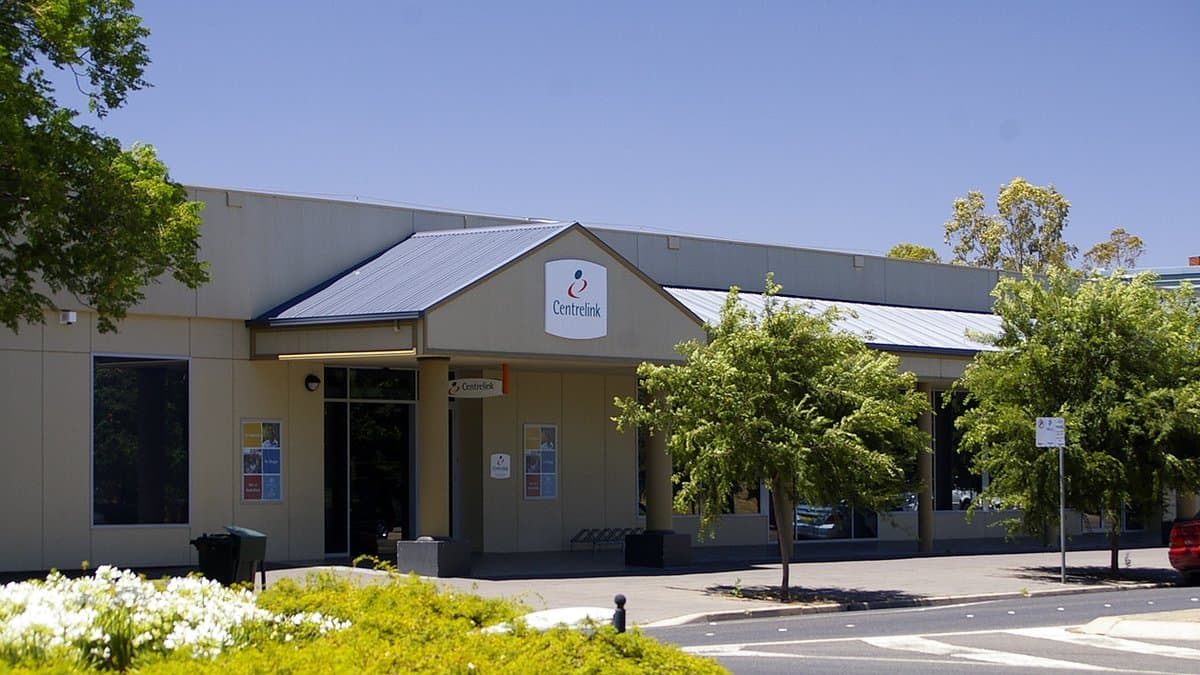A low government service center with Centrelink signage above its entrance, seen from the street on a sunny day, with trees and a parked car alongside.