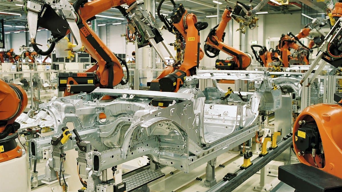 Rows of orange industrial robot arms working on a bare steel car body on an automated production line.