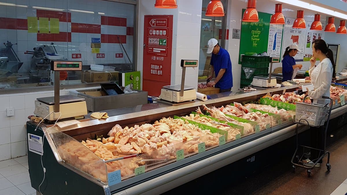 Trays of poultry and cuts of meat in a refrigerated display case at a supermarket meat counter, with hanging lamps above and staff working behind the counter.