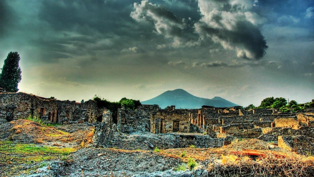 The excavated stone ruins of Pompeii in the foreground, with Mount Vesuvius rising behind them under a dark, clouded sky.