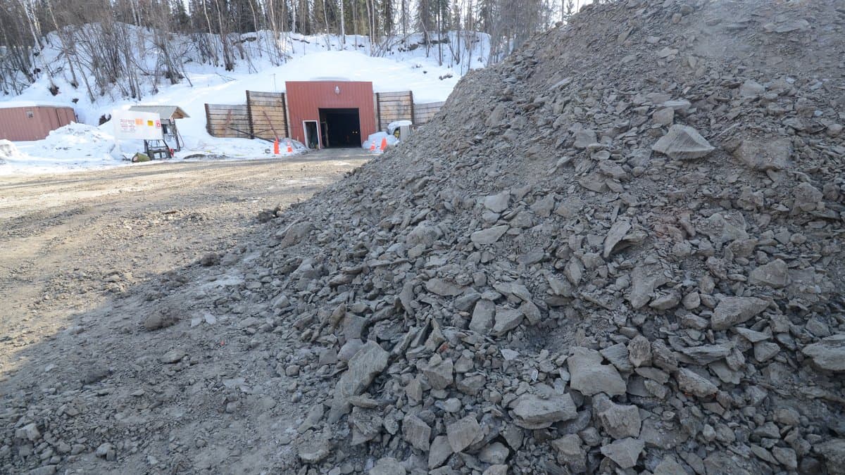 A tunnel portal set into a snowy hillside in a boreal forest, with a large pile of excavated gray frozen soil in the foreground.