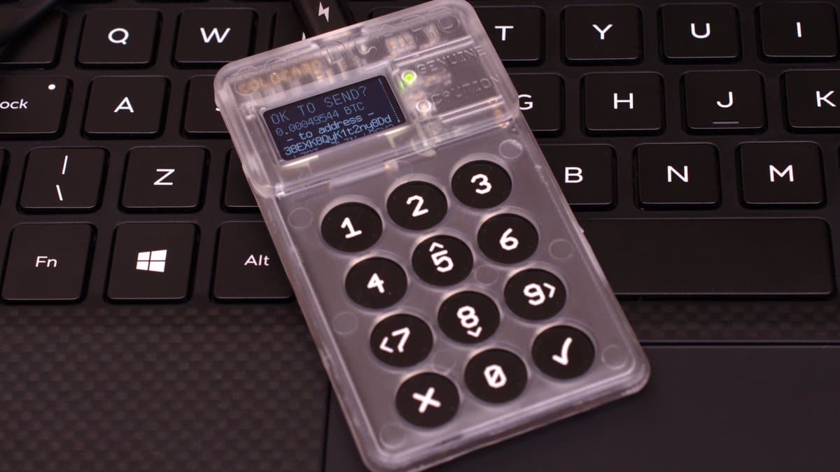 A translucent plastic hardware wallet with a numeric keypad and a small lit display, lying on a black laptop keyboard.