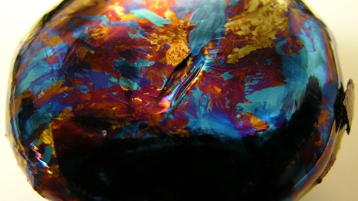 A rounded lump of hafnium metal whose surface oxide layer shows bands of blue, orange and red interference color.