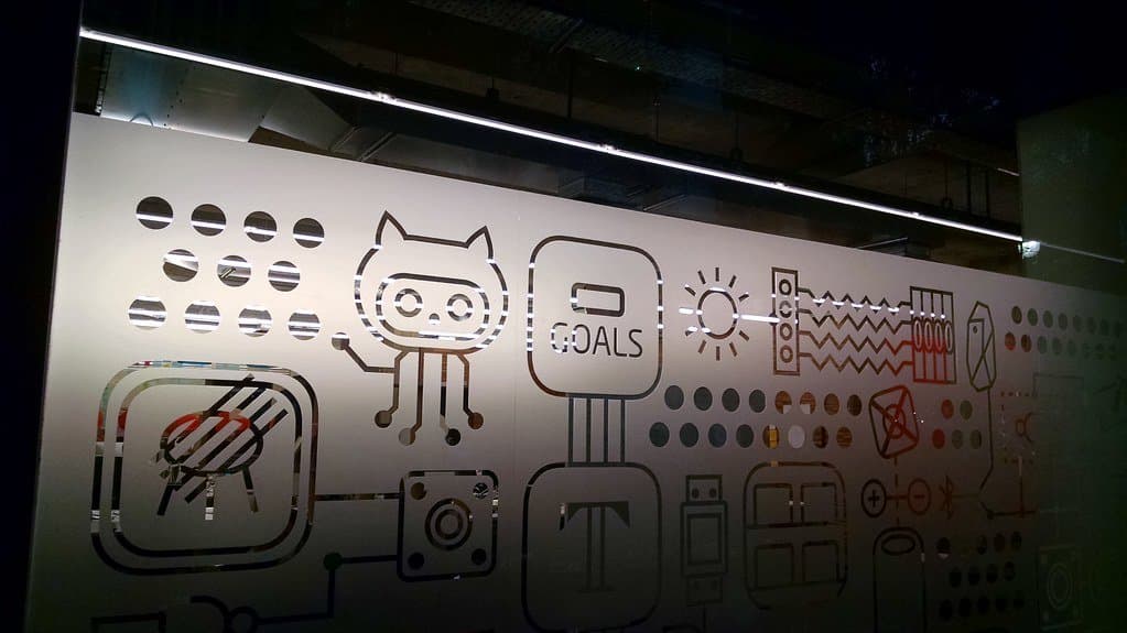 A backlit glass panel etched with GitHub's Octocat mascot and rows of small technical icons, seen inside a building at night.