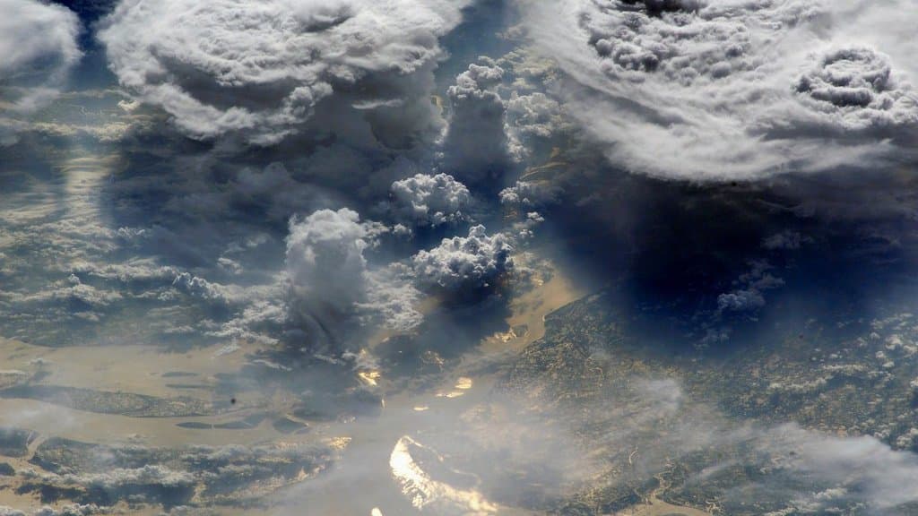 Towering monsoon storm clouds photographed from orbit over the river channels of the Ganges-Brahmaputra delta in Bangladesh, with sunlight glinting off the water below.