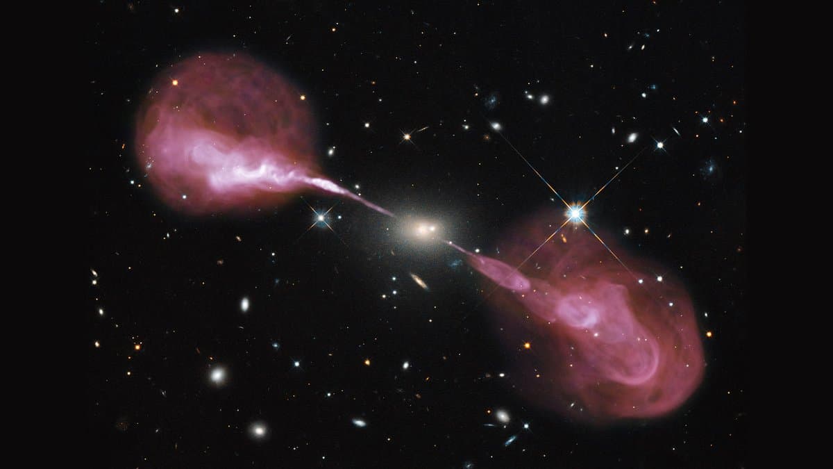 The elliptical galaxy Hercules A at center, with two pink jets from its core extending to opposite sides into large lobes, set against a field of distant galaxies.