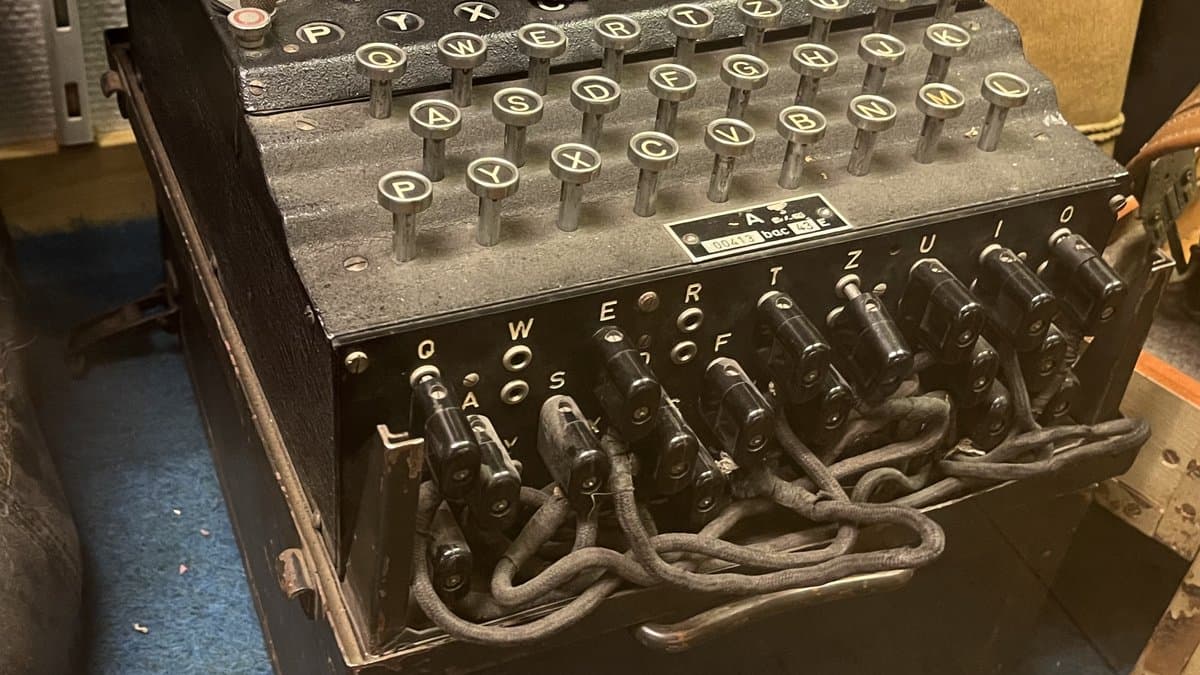 A black three-rotor Enigma cipher machine seen from above, showing its QWERTZ keyboard and the front plugboard with patch cables plugged into lettered sockets.