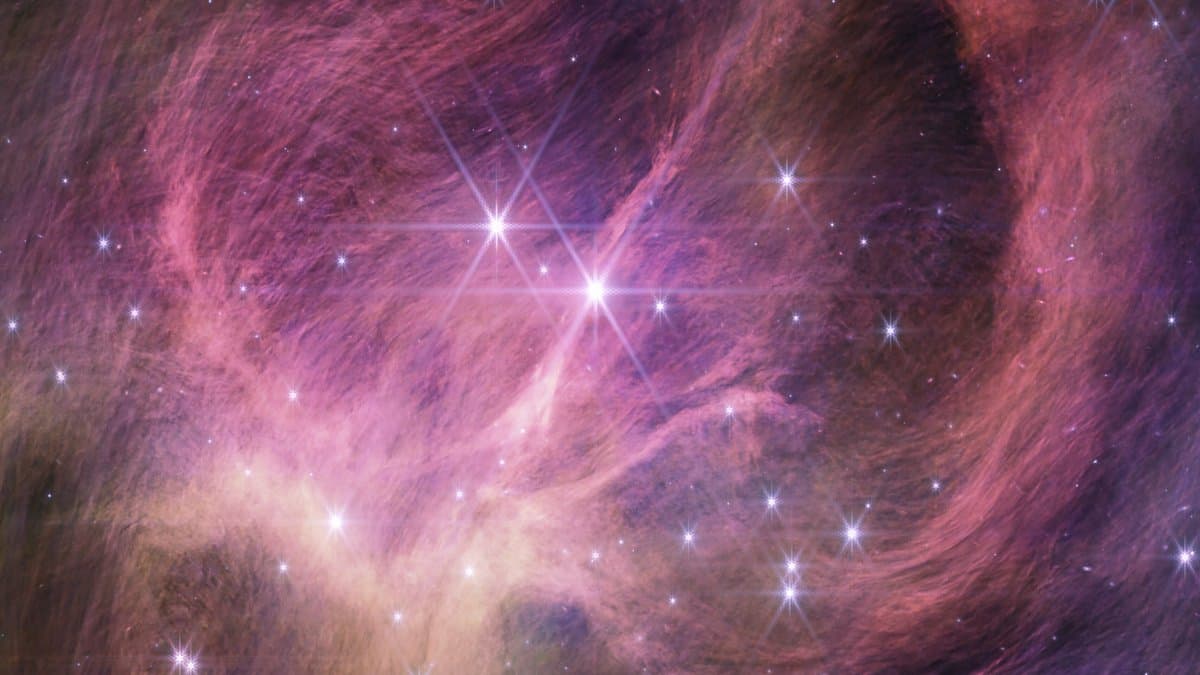 A dense field of stars behind wispy pink and white clouds of dust and gas, imaged in near-infrared light by the James Webb Space Telescope.