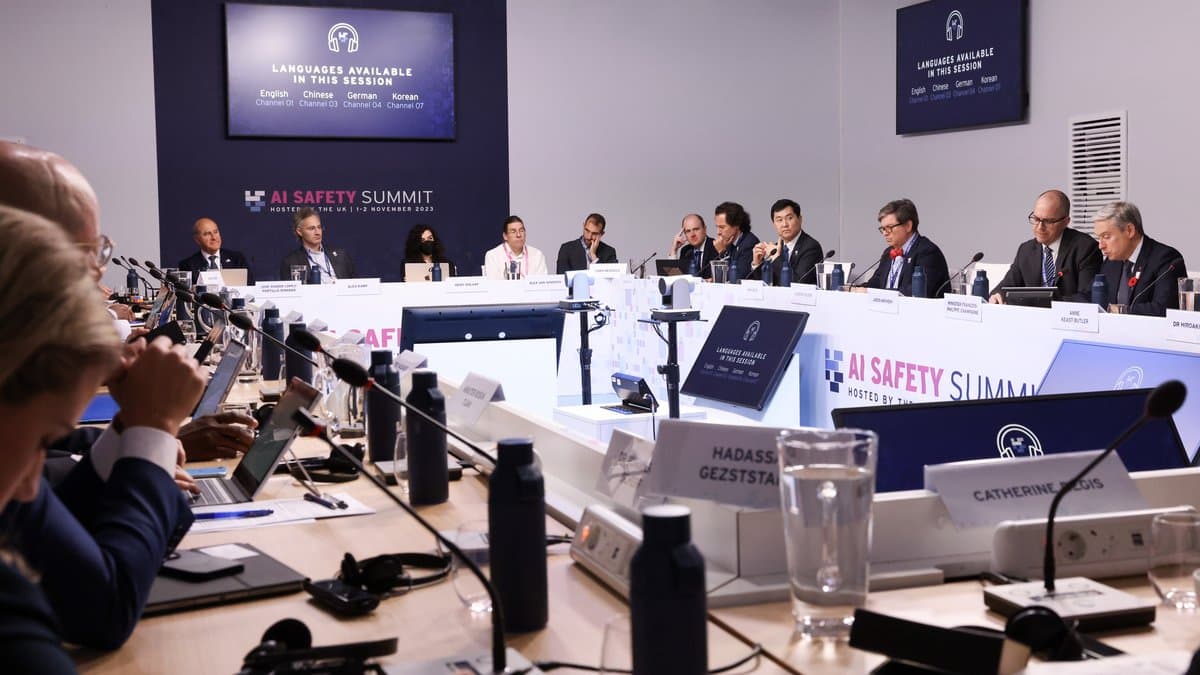 Delegates in dark suits seated around a large square conference table, with translation screens and AI Safety Summit signage on the wall behind them.
