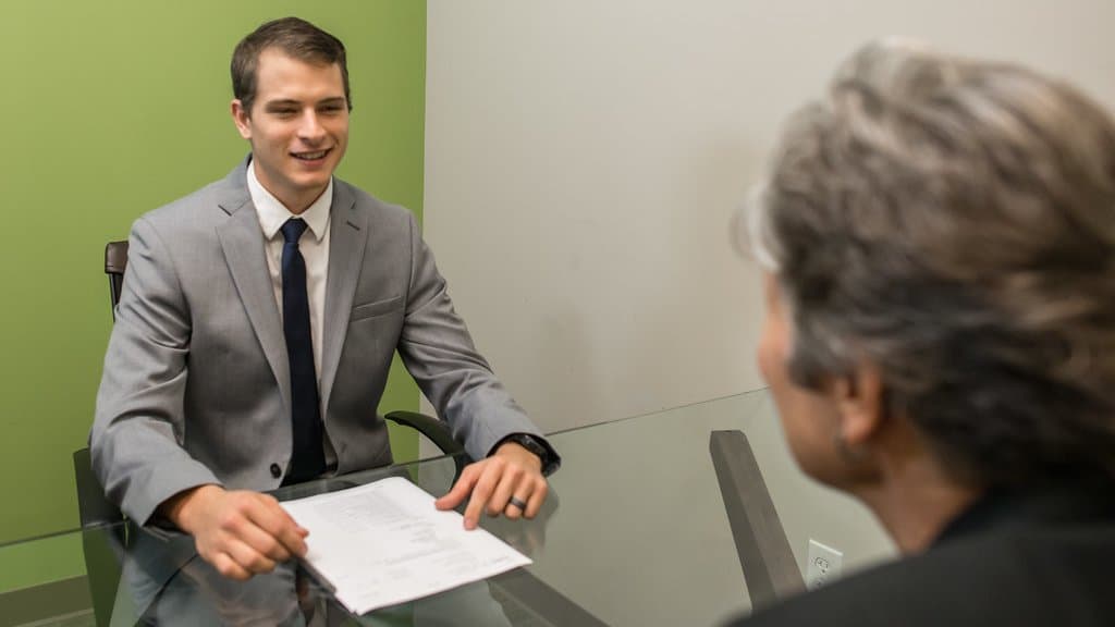 A job applicant in a suit hands a printed resume across a glass table to an interviewer.