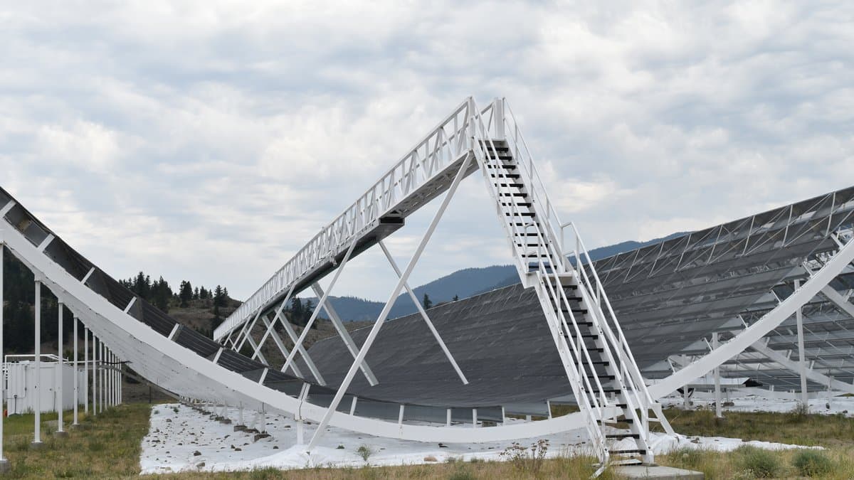 A long white cylindrical radio telescope reflector with a dark wire-mesh surface, with forested hills behind it.