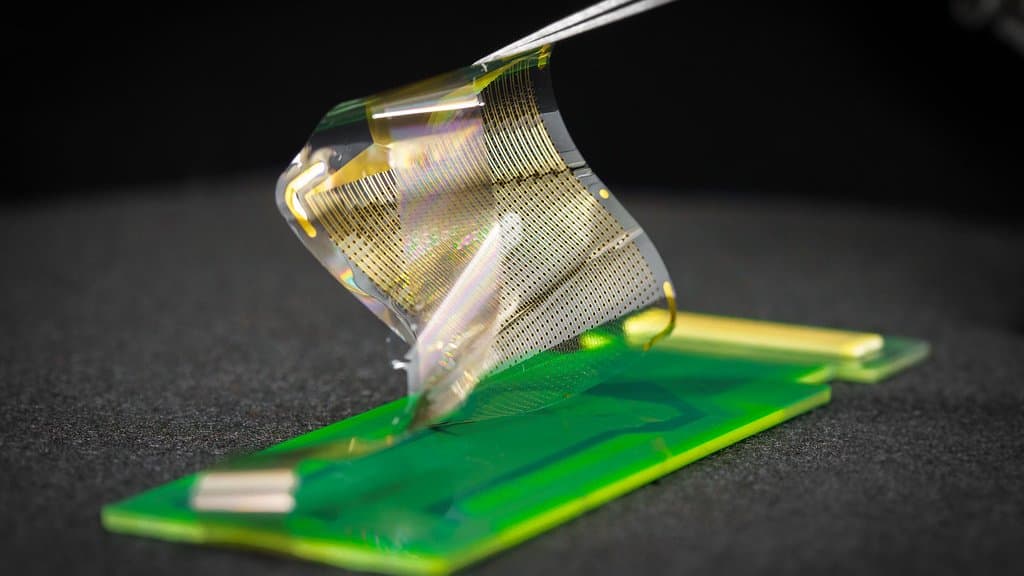A transparent thin-film electrode array with hundreds of fine gold contacts, lifted with tweezers above a green connector board on a dark benchtop.