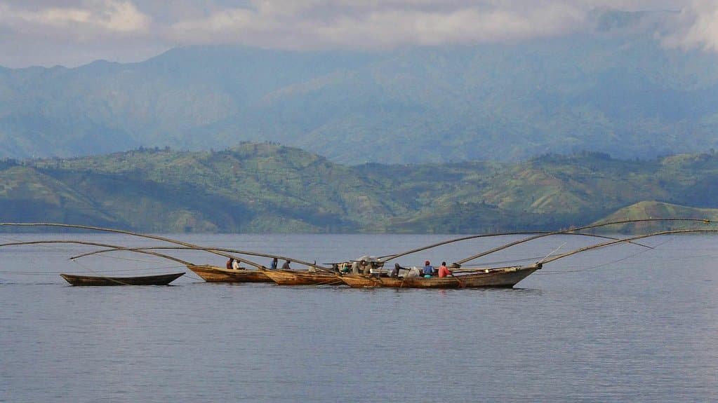Long wooden fishing canoes with arched poles rest on the calm water of Lake Kivu, with green hills and a ridge of mountains behind them.