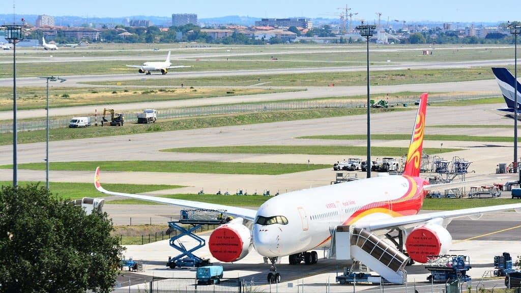A white and red Airbus A350 passenger jet parked on an apron at Toulouse-Blagnac airport in France, with maintenance stands beside it, runways behind and the city on the horizon.